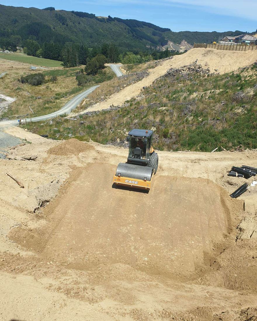 Roller compacting a fill platform on a hillside earthworks site, with a road winding behind