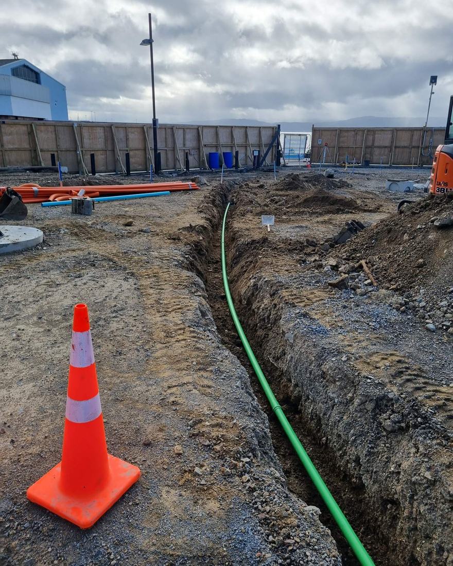 Open trench with a green service duct laid along it and a road cone at the edge