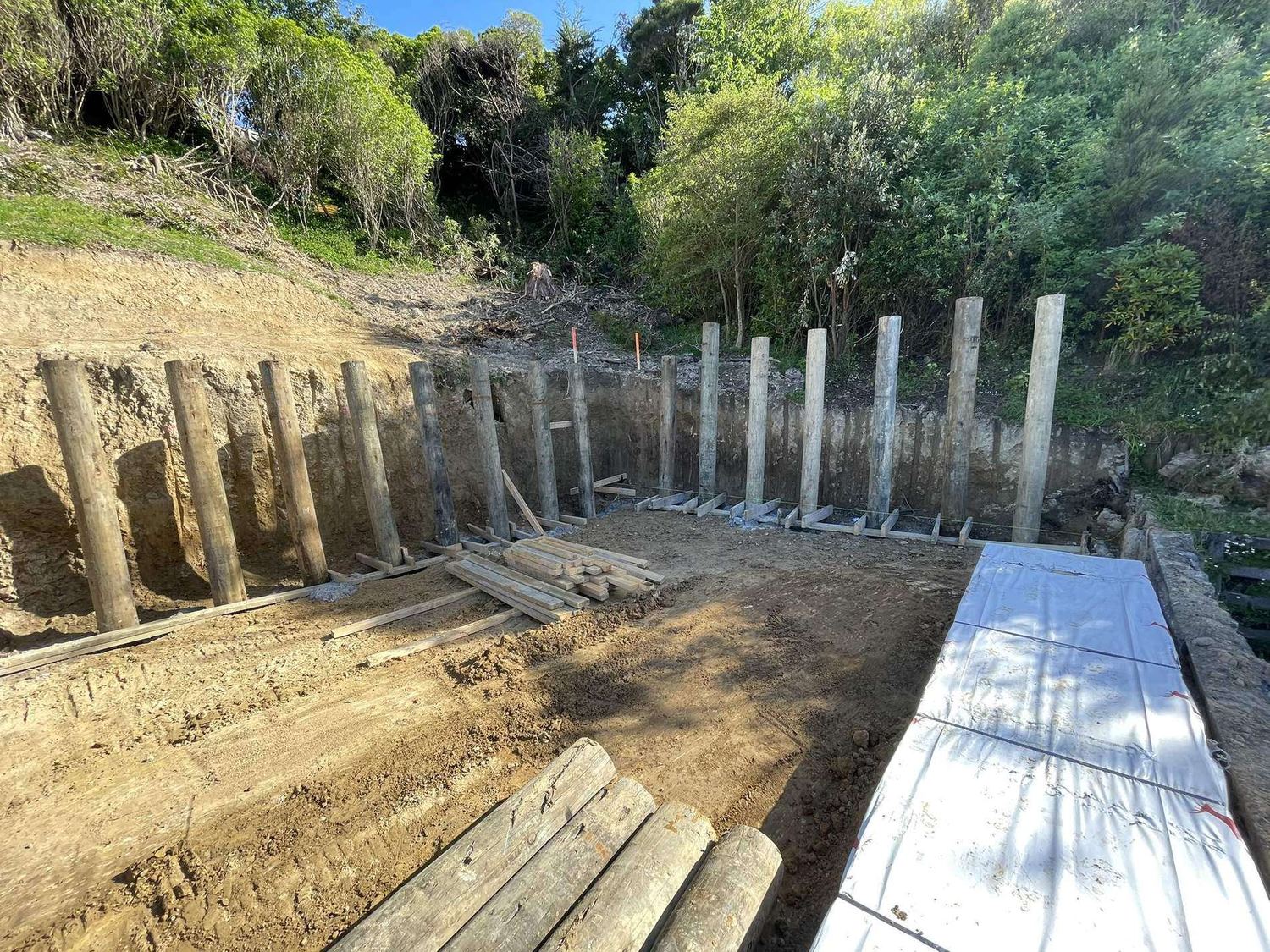 Timber poles set for a retaining wall against a bush bank, with lagging boards ready to fit