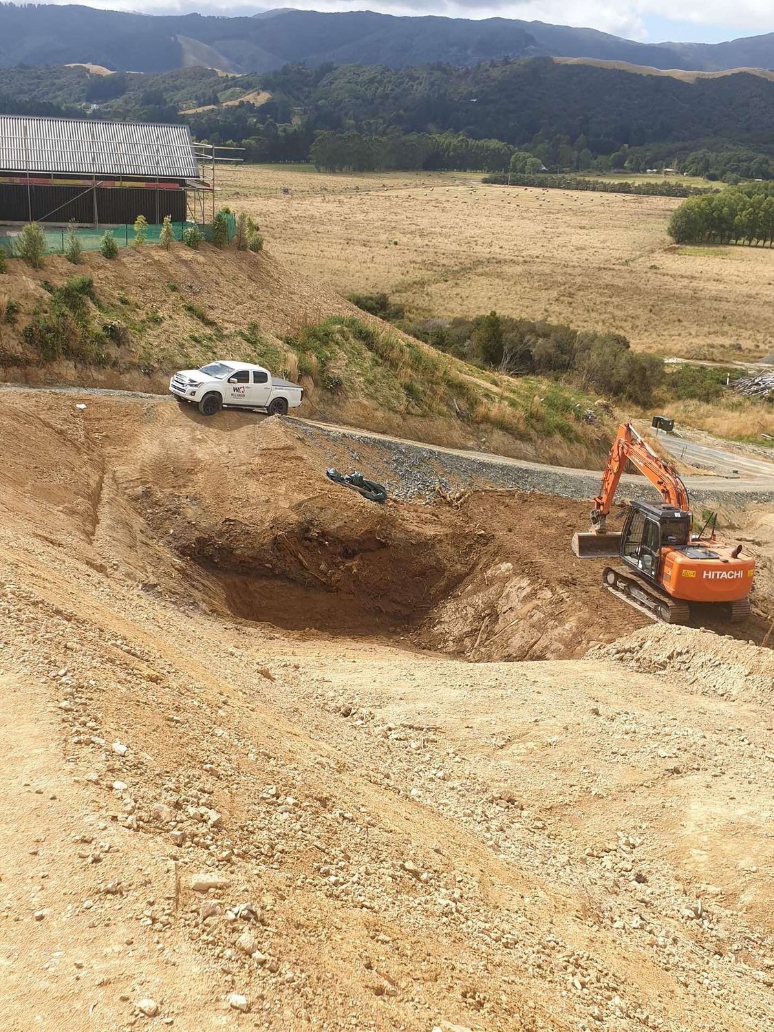 Excavator digging out a large hole on a rural site, with a ute and farm buildings above