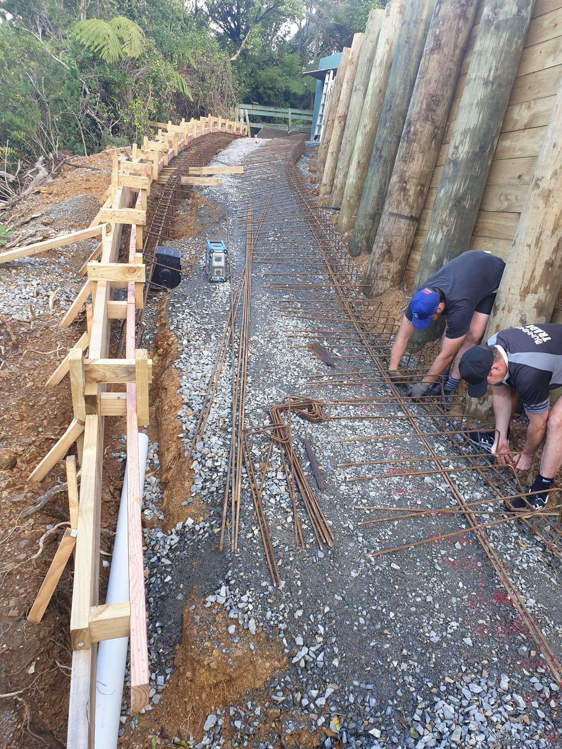 Workers tying reinforcing steel in timber boxing for a footing in front of a pole wall